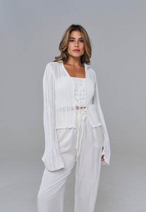Femme portant un cardigan blanc en maille par-dessus un crop top blanc avec cordons, associé à un pantalon ample en satin blanc, debout devant un fond gris.