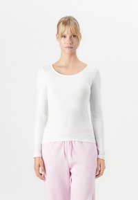 Young woman with blonde hair in a ponytail wearing a white long-sleeve ribbed top and light pink pants, standing against a plain white background.