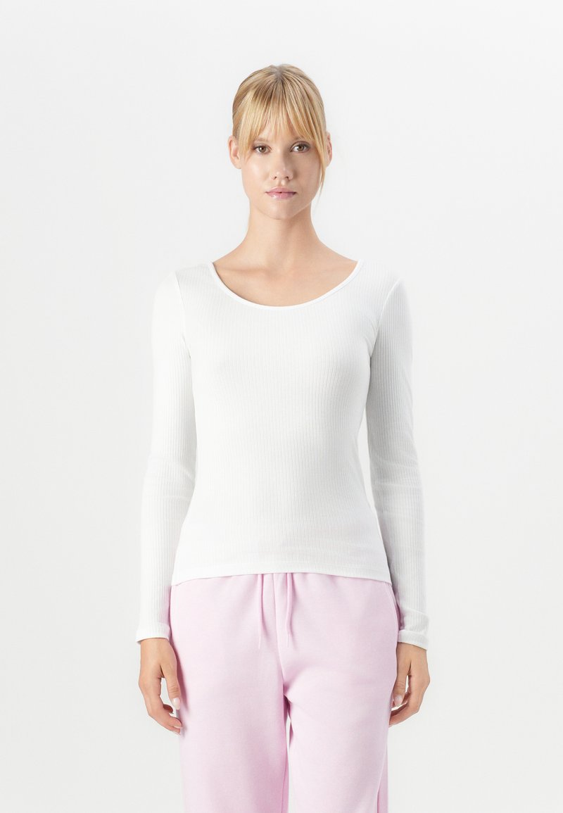 Young woman with blonde hair in a ponytail wearing a white long-sleeve ribbed top and light pink pants, standing against a plain white background.