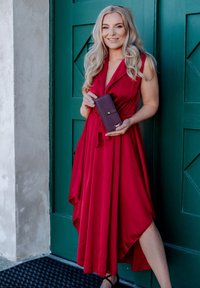 Red sleeveless dress with a tie waist, flowing skirt, paired with a textured purple wallet featuring a gold accent. Background: green door.