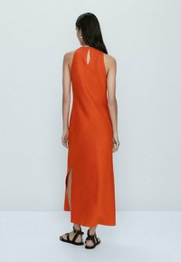 Długa, bezrękawna pomarańczowa sukienka z gładkiego materiału, z detalem w kształcie dziurki na plecach i rozcięciem po boku. Noszona z czarnymi sandałami.
