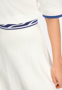 Weißer plissierter Rock und weißes Kurzarmoberteil mit blauen und rosa gestreiften Bündchen sowie tailliertem blauen Muster auf dem Oberkörper und Arm einer Person.