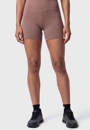 Person, die mauvefarbene, hoch taillierte Biker-Shorts, schwarze Champion-Socken und schwarze Sportschuhe trägt, steht auf einem schlichten hellen Hintergrund.