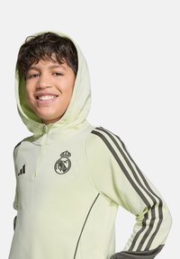 Sweatshirt à capuche jaune avec des rayures et des détails noirs, arborant le logo du Real Madrid sur la poitrine et un col zippé.