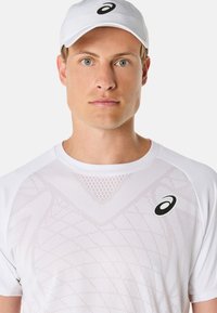 Bílý sportovní trikot s texturovaným síťovaným vzorem, se černým logem. V kombinaci s odpovídající bílou čepicí. Hladká tkanina s přiléhavým střihem.