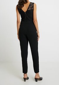 Mono negro con un diseño ajustado, que cuenta con un espalda de encaje, corte sin mangas y pantalones ajustados, combinado con mule negros de tacón alto.