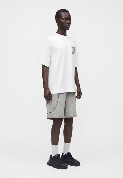 Άνδρας όρθιος με λευκό φαρδύ T-shirt με λογότυπο, ανοιχτόγριες βερμούδες με μαύρα ραφές, λευκές κάλτσες και μαύρα αθλητικά παπούτσια σε λευκό φόντο.