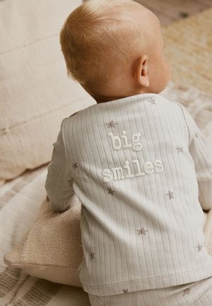 Dieťa oblečené v bielom rebrovanom outfite so sivými hviezdami, plazí sa po béžovej deke, text "veľké úsmevy" na zadnej strane outfitu.