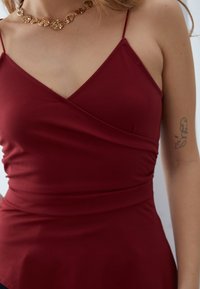 Bordo bezrukavni top s wrap dizajnom, izrađen od glatkog materijala. Ima tanke naramenice i nabran detalj na struku.