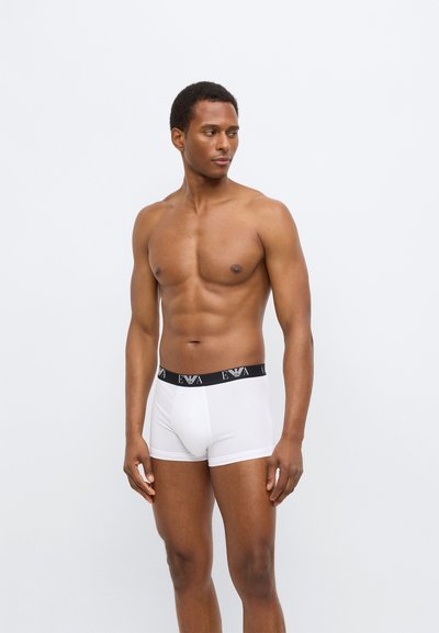 Άσπρα βαμβακερά αντρικά μπόξερ με μαύρη μέση που φέρει λογότυπο. Απαλή υφή, εφαρμοστή σχεδίαση και μήκος μέχρι το μέσο του μηρού.