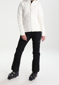 Doudoune blanche à capuche, avec un motif matelassé diagonal et une fermeture éclair à l'avant. Pantalon de ski noir et bottes complètent la tenue.