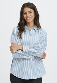 Smiling woman with long dark hair wears a blue and white vertical striped button-up shirt, standing with arms crossed on a white background.