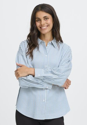Femme souriante avec de longs cheveux noirs portant une chemise à rayures verticales bleues et blanches, se tenant les bras croisés sur un fond blanc.