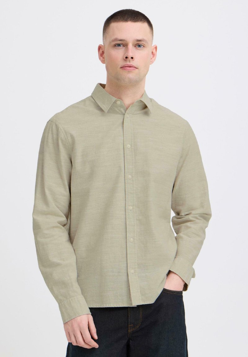 Homme portant une chemise beige à manches longues avec boutons et un pantalon foncé à poches, debout devant un fond blanc uni.