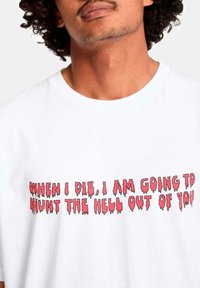 Hombre con cabello rizado vistiendo una camiseta blanca con texto rojo: "CUANDO MUERA, TE VENGARÉ EN EL INFIERNO."