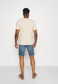 Camiseta de algodón beige con mangas cortas, combinada con pantalones cortos de mezclilla azules, detalles de bolsillo visibles y zapatos marrones de tipo slip-on.