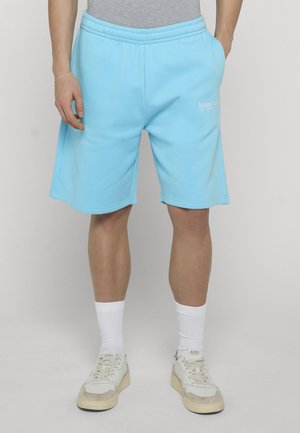 Person, die hellblaue lässige Shorts, weiße Socken und weiße Turnschuhe trägt, steht mit den Händen in den Taschen vor einem schlichten Hintergrund.