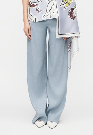 Hellblaue Hosen mit weitem Bein, kombiniert mit weißen Spitzschuhen, getragen zu einem Oberteil mit buntem Engel-Print und asymmetrischem Saum.