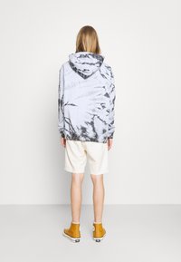 Persona con cabello largo que lleva una sudadera de tie-dye en blanco y negro, pantalones cortos beiges y zapatillas altas mostaza de espalda frente al espectador.