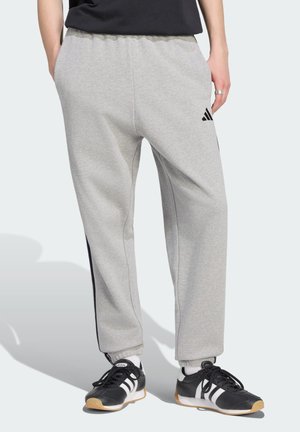 Person iført lysegrå joggingbukser med sort logo og sort-hvide sneakers med gummisåler, stående mod en ensfarvet baggrund.