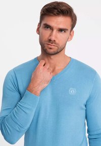Maglione aderente azzurro chiaro con maniche lunghe e scollatura a V, dotato di un logo discreto al centro. Texture morbida e design pulito.