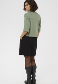 Zöld, rövid ujjú pulóver laza szabással, fekete szoknyával és térdig érő fekete bőr csizmával párosítva. Sima textúra és hétköznapi dizájn.