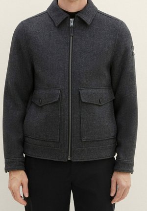 Mann trägt eine dunkelgraue Wolljacke mit Frontreißverschluss und zwei großen, mit Knöpfen verschlossenen Klappentaschen, steht vor einem schlichten Hintergrund.