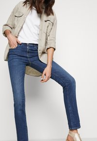 Blå skinny jeans i denim, tillsammans med en ljusbeige skjorta med knappar och en vit t-shirt. Skorna är remmade och i metallisk guld.