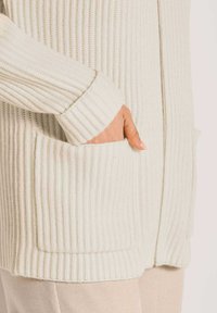 Hand teilweise in der Tasche eines cremefarbene, gerippten Strickjacke mit Reißverschluss, die über beigen Hosen getragen wird.