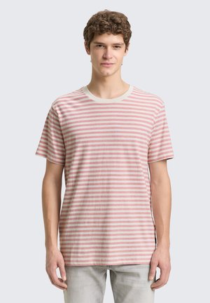 Junger Mann mit kurzen, lockigen Haaren, der ein kurzärmeliges T-Shirt mit hellrosa-weißen Streifen und hellgraue Hosen trägt, vor einem einfarbigen Hintergrund stehend.