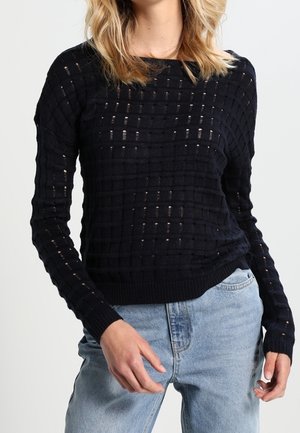 Vrouw draagt een zwarte, lange mouw, gebreide trui met een rasterpatroon en lichtblauwe, hoog getailleerde jeans, staand tegen een effen achtergrond.