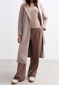 Beige lång kappa över en ljusbeige tröja, matchad med bruna veckade byxor och krämfärgade sneakers med bruna detaljer. Släta texturer, modern passform.