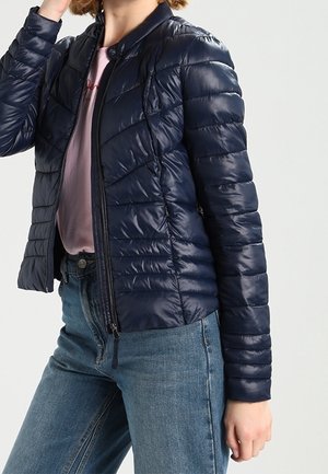 Person trägt eine marineblaue gesteppte Jacke über einem rosa Hemd und blauen Jeans, mit einer Hand in der Nähe des Gesichts.