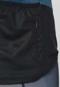 Musta urheilu paita, jossa on teksturoitu materiaali, joustava vyötärö ja sivussa vetoketjullinen tasku. Suunnittelussa on sininen yksityiskohta.