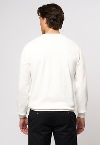 Sweatshirt blanc à manches longues et poignets côtelés, avec un col rond et une texture douce. Porté avec un pantalon foncé, affichant un design simple.