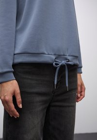Jasnoszaro-niebieski sweatshirt o gładkiej teksturze, z regulowanym sznurkiem w talii i luźnym fasonie, połączony z ciemnymi dżinsami.