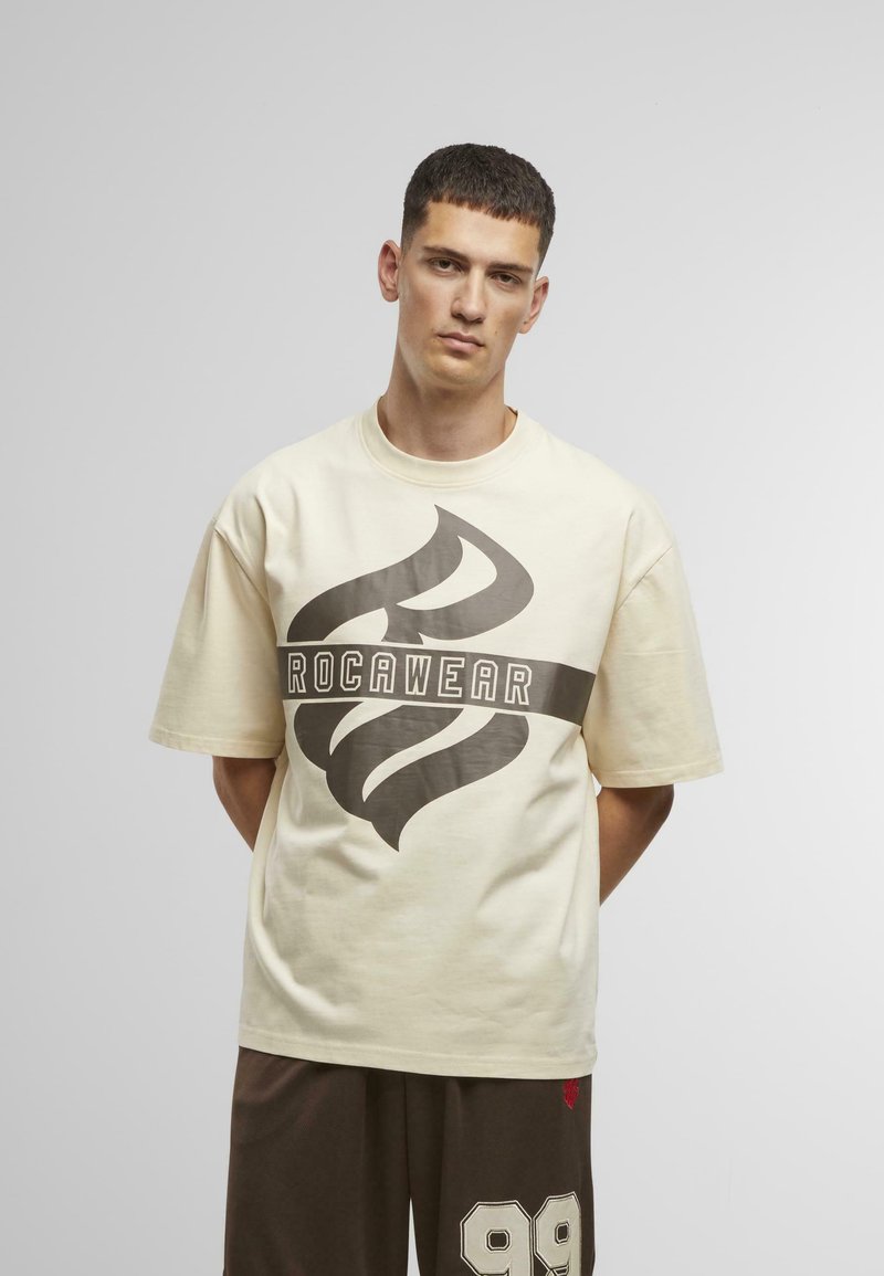 T-shirt couleur crème avec un logo ROC-A-WEAR brun audacieux, à manches courtes et à coupe décontractée, associé à un short marron foncé avec le numéro 99.