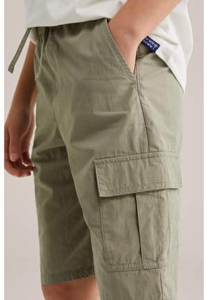 Copil purtând pantaloni scurți cargo verde deschis cu buzunar lateral mare și tricou alb, cu mâna în buzunarul pantalonilor, pe fundal neutru.