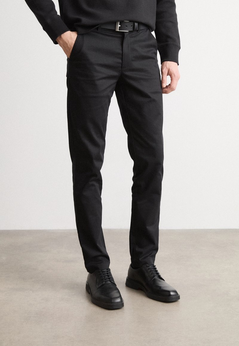 Homme portant un pantalon slim noir, une ceinture noire, une chemise noire à manches longues et des chaussures de ville noires, debout sur un sol gris.