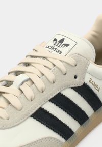 Κοντινό πλάνο του αθλητικού παπουτσιού Adidas Samba με μπεζ κορδόνια, μαύρες ρίγες και σουέντ λεπτομέρειες σε λευκό φόντο.