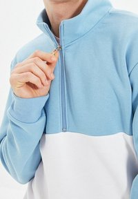 Osoba zapínající světle modrý a bílý svetr na půl zip, ruka držící tahák zipu blízko límce.