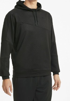 Mann trägt schwarzen Pullover-Hoodie mit geometrischem Muster und schwarze Hose, steht vor weißem Hintergrund.