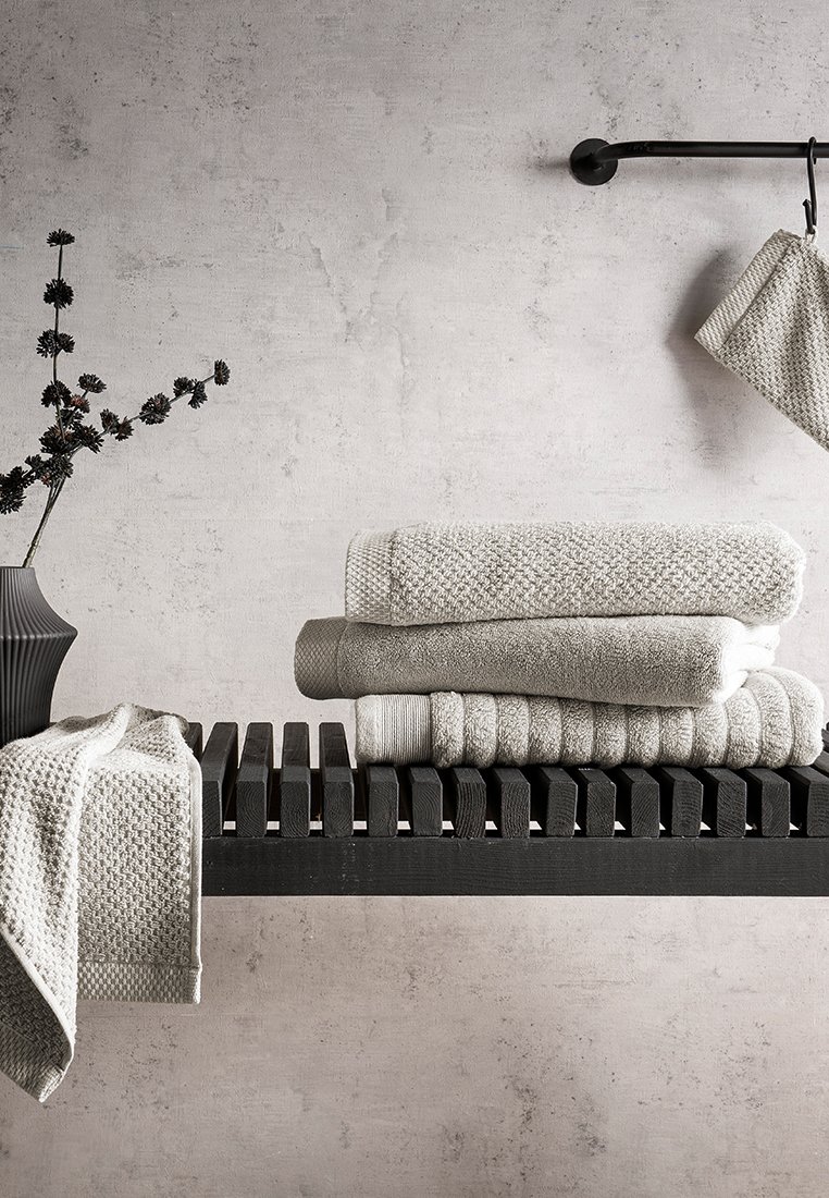 Schwarzes Holzregal, das drei übereinander gestapelte graue Handtücher mit unterschiedlichen Texturen zeigt, während ein graues, gemustertes Handtuch über die Kante hängt. Neben dem Regal steht eine schwarze Vase mit dunklen Blumen.