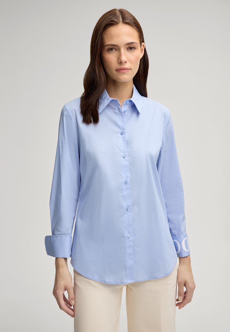 Camicia a maniche lunghe blu chiaro con chiusura a bottoni e orlo curvo. I polsini presentano un motivo circolare. Tessuto dalla texture liscia.