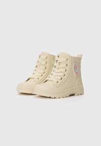 Botas altas beige de material de goma, con punta redonda, suela gruesa, diseño de cordones y un parche en forma de corazón con una ilustración de un unicornio.
