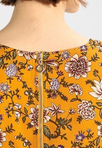 Záda osoby s viditelným krkem a horní částí zad oblečenou do hořčicově žlutých květinových šatů s viditelným stříbrným zipem a kulatou táhlou šňůrkou.