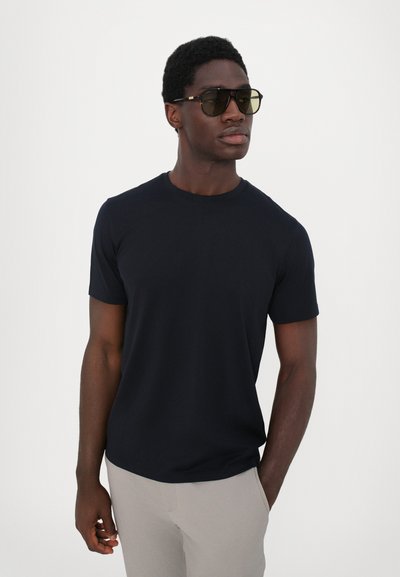 Schwarzes T-Shirt mit kurzen Ärmeln aus glattem Stoff, kombiniert mit grauen Hosen. Das Modell trägt dunkle Sonnenbrillen mit einem geometrischen Rahmen-Design.