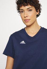 T-shirt en coton bleu marine avec un design à col en V. Il présente un logo Adidas brodé en blanc sur la poitrine gauche. Manches courtes, texture douce.