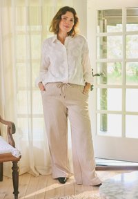 Femme debout à l'intérieur près d'une porte en verre, vêtue d'une chemise blanche boutonnée et d'un pantalon beige à jambes larges, les mains dans les poches.