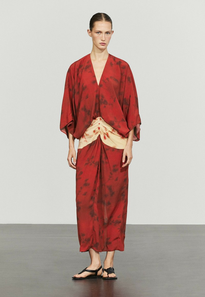 Woman standing wearing a loose red and beige tie-dye dress with wide sleeves and black flat sandals against plain background.
